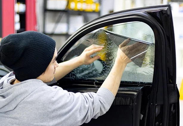A technician wearing a black beanie carefully applying window tint film to the side window of a dark vehicle using a squeegee while water solution helps position the film. The installer smooths the tint across the glass during a professional window tint installation service at Inch X Inch in Sarasota Florida located at 640 Apex Rd Sarasota FL.
