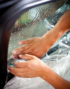 Close up view of hands pressing and smoothing window tint film along the edge of a vehicle window while installation solution is visible on the glass. The technician carefully aligns the film to ensure a clean finish during a window tint service at Inch X Inch in Sarasota Florida.